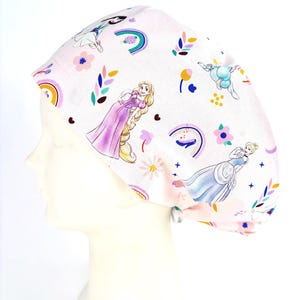 May include: A light pink scrub cap featuring a repeating pattern of cartoon princesses, colourful rainbows, small flowers, and leaves. The fabric has a soft pastel background with various illustrated characters in dresses, including figures with long blonde hair.