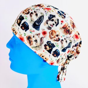 May include: A white surgical cap patterned with multiple dog breeds and red floral designs. The cap is tied at the back and is shown on a blue mannequin head. The fabric has a soft, slightly textured appearance.