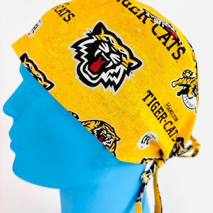 May include: Yellow scrub cap featuring a tiger logo and the words "TIGER-CATS" and "HAMILTON FOOTBALL". The cap has yellow ties at the back. The cap is patterned.