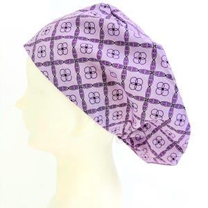 May include: A light purple scrub cap with a repeating floral pattern in dark purple and black. The cap is on a mannequin head, showcasing the design and fit. The cap is designed to cover the hair.