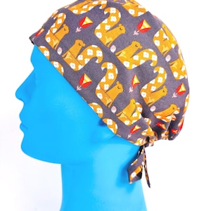 May include: A patterned scrub cap featuring a repeating design of yellow squirrels with white flower accents on a dark gray background. The cap has a tie closure at the back and is shown on a blue mannequin head.