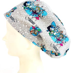 May include: A gray surgical scrub cap featuring a pattern of cartoon characters and white stars. The cap has a blue trim and the words "The Jetsons" in a cursive font. The cartoon characters are in blue ovals.