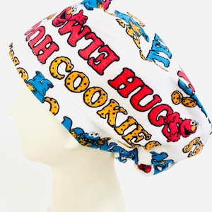 May include: White scrub hat with a blue and red Sesame Street theme. The hat features Cookie Monster and Elmo with the text "Me Want Cookie" and "Big Hugs".