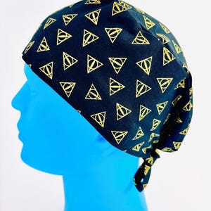 May include: A black surgical scrub cap featuring a repeating gold-colored Deathly Hallows symbol pattern. The cap is tied at the back and is designed to cover the head and hair. The cap is shown on a blue mannequin head.