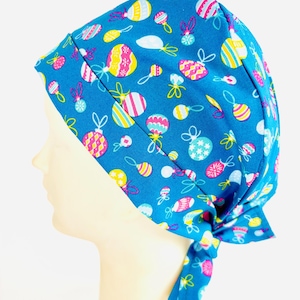 May include: A blue scrub cap with a pattern of colourful Easter egg and rattle designs. The cap is tied at the back and is made of a soft, lightweight fabric. The Easter-themed cap is designed for medical professionals or anyone needing hair protection.