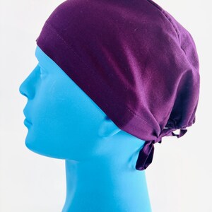 May include: A purple scrub hat with a tie at the back. The hat is made of a soft, comfortable fabric.