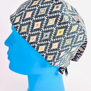 May include: A patterned surgical scrub cap with a repeating diamond design in black, white, yellow, orange, pink, and blue. The cap is tied at the back and is shown on a blue mannequin head.