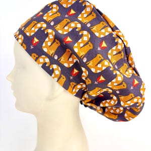 May include: A patterned scrub cap with a repeating design of orange, stylized animals, white floral accents, and red kite shapes on a dark purple background. The cap is made of fabric and is designed to be worn on the head.