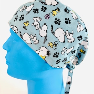 May include: A light blue surgical scrub cap featuring a repeating pattern of Snoopy, Woodstock, paw prints, and flowers. The cap is tied at the back and is made of a soft, lightweight fabric. The design is playful and suitable for medical professionals.