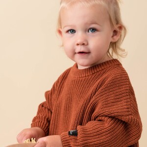 May include: A young child wearing a rust-colored knitted sweater with a small black logo on the sleeve. The child is holding a wooden comb with a small wooden brush attached to it.