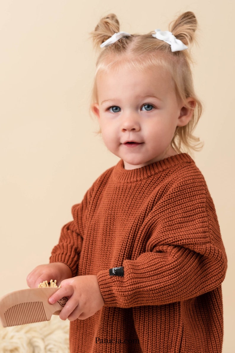 May include: A young child wearing a rust-colored knitted sweater with a small black logo on the sleeve. The child is holding a wooden comb with a small wooden brush attached to it.