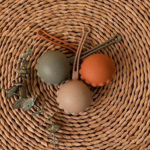 May include: Three silicone teethers in the shape of a bear with different colored faces: green, brown, and orange. The teethers are on a woven brown surface.