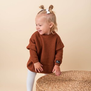 May include: A young child wearing a brown knitted sweater with white bows in their hair, standing on a woven round stool.