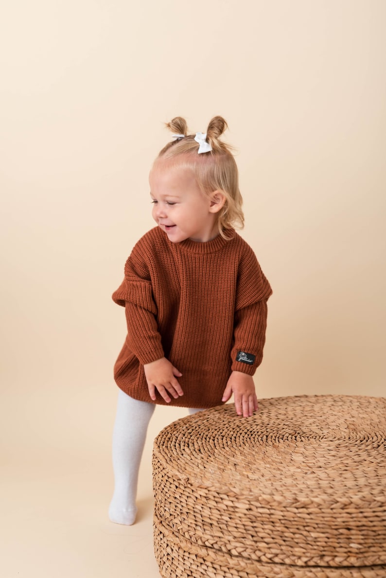 May include: A young child wearing a brown knitted sweater with white bows in their hair, standing on a woven round stool.