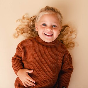 May include: A young girl with long, curly blonde hair is wearing a brown knitted sweater. She is lying on her back and smiling at the camera.