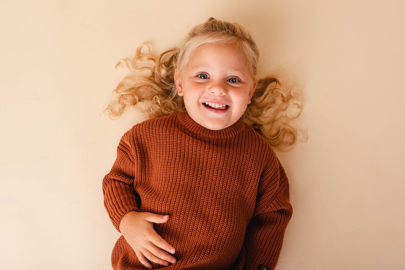 May include: A young girl with long, curly blonde hair is wearing a brown knitted sweater. She is lying on her back and smiling at the camera.
