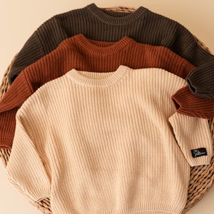 May include: Three knitted sweaters in a basket. The sweaters are in shades of brown, rust, and cream.