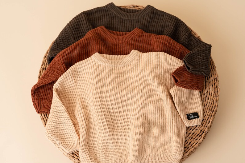 May include: Three knitted sweaters in a basket. The sweaters are in shades of brown, rust, and cream.