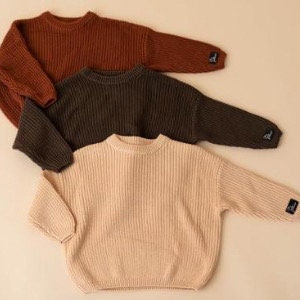 May include: Three knitted sweaters in rust, dark green, and beige. The sweaters have a ribbed texture and a relaxed fit.