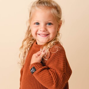 May include: A young girl with long blonde hair smiles while wearing a rust-colored knitted sweater.