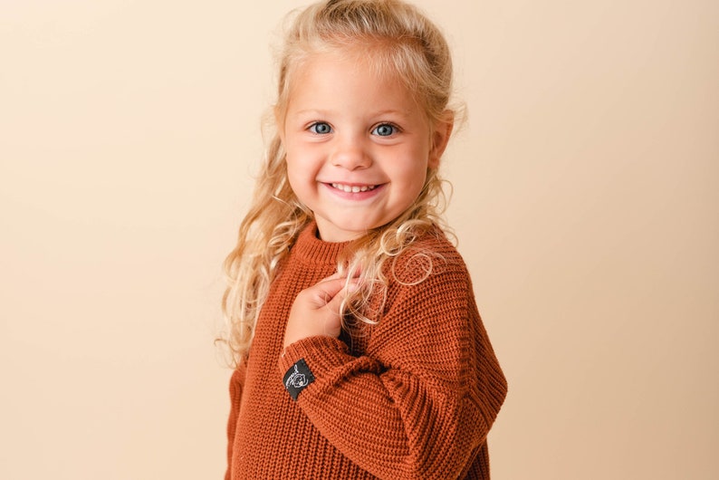 May include: A young girl with long blonde hair smiles while wearing a rust-colored knitted sweater.