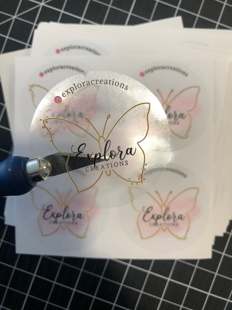 May include: Clear sticker with a gold butterfly design and the text "Explora Creations". The butterfly has a pink watercolor wash.