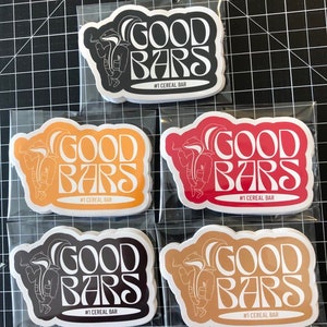 May include: Five stickers with a white outline of a cartoon badger wearing a jacket and holding a cigarette. The text "GOOD BARS" is in a retro font and the text "#1 CEREAL BAR" is below the badger. The stickers are on a white background and are in different colors: black, orange, red, brown, and beige.