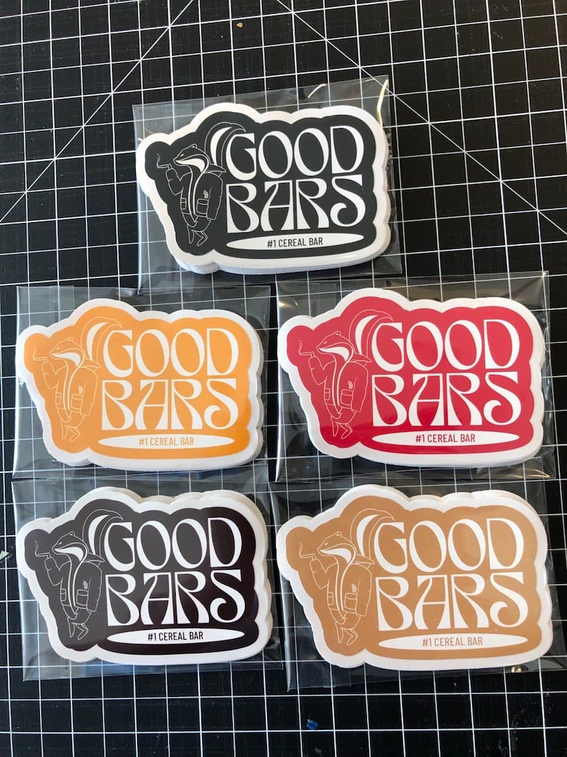 May include: Five stickers with a white outline of a cartoon badger wearing a jacket and holding a cigarette. The text "GOOD BARS" is in a retro font and the text "#1 CEREAL BAR" is below the badger. The stickers are on a white background and are in different colors: black, orange, red, brown, and beige.