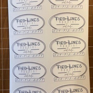 Puede incluir: Diez pegatinas ovaladas blancas con un contorno negro. Cada pegatina tiene el texto "TIED LINES Apparel and more..." y un n&uacute;mero de tel&eacute;fono "504-912-6588".