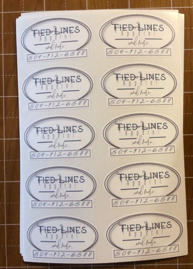 Puede incluir: Diez pegatinas ovaladas blancas con un contorno negro. Cada pegatina tiene el texto "TIED LINES Apparel and more..." y un n&uacute;mero de tel&eacute;fono "504-912-6588".