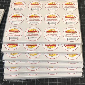 May include: A stack of white sheets of paper with red and yellow circular stickers. Each sticker has a red border and a yellow interior with a cartoon image of a train and the text "Old Fashioned Banana Pudding" and a phone number.