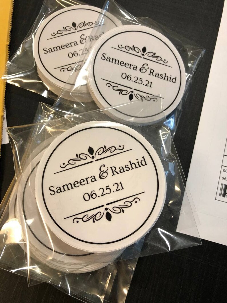May include: A stack of white round stickers with black text and decorative borders. The text reads "Sameera & Rashid 06.25.21".