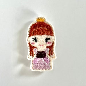 May include: Embroidered patch featuring a cartoon princess with red hair, a gold crown, and a pink dress. The patch has a white border and is designed with detailed stitching. The princess has a sweet expression.