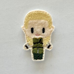 May include: Embroidered patch featuring a cartoon-style figure with blonde hair and a green outfit. The patch has black eyes and a small pink mouth. The figure's arms have silver and black details. The patch is on a white background.