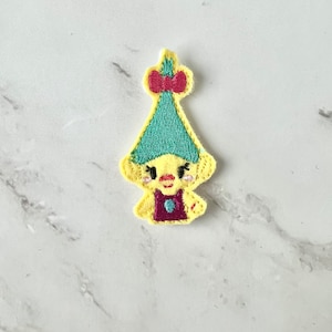 May include: Yellow felt embroidered gnome with a teal hat and a pink bow. The gnome is wearing a purple dress with a teal gem.