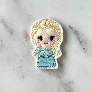 Puede incluir: Parche de fieltro bordado de una princesa de dibujos animados con cabello rubio, vistiendo un vestido azul.