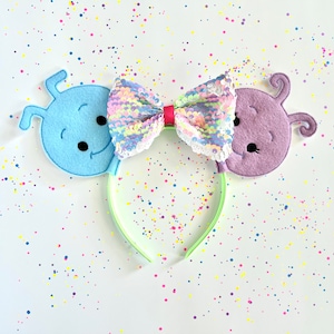 May include: A pair of light blue and purple felt character ears with a pink and iridescent sequin bow on a green headband. The ears are shaped like aliens with large eyes and smiles. The background is white with colorful confetti.