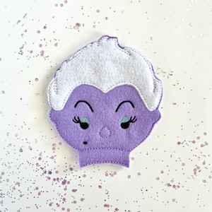 May include: A felt Ursula the Sea Witch face in shades of purple, white, and black. The character has a white wig, black eyelashes, and a beauty mark. The felt is stitched together with white thread. The background is white with scattered glitter.