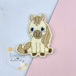 Puede incluir: Un parche bordado de fieltro beige con un caballo de dibujos animados con melena y cola marrones. El caballo tiene la nariz marrón, los ojos negros y las orejas rosas.