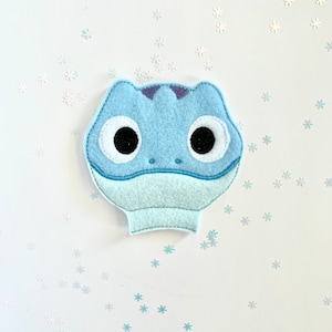 May include: A light blue felt applique in the shape of a cartoon lizard's head. The lizard has large black eyes with white circles, a light blue snout, and a purple crest. The background is white with scattered snowflake confetti.