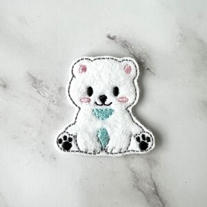 May include: White felt applique of a polar bear with black paws and a light blue heart on its chest. The bear has pink cheeks and black eyes.