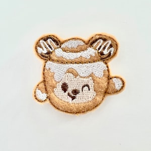 Puede incluir: Un parche bordado con un oso de dibujos animados con una expresión feliz. El oso es marrón y blanco, con una raya blanca en la cabeza y lleva un sombrero blanco.