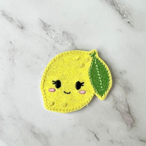Può includere: Un limone di feltro giallo con una foglia di feltro verde e un viso sorridente. Il limone ha le guance rosa e gli occhi neri.