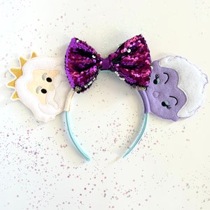Peut inclure: Un serre-tête avec un nœud en sequins violets et deux oreilles en feutre représentant des personnages. L'oreille gauche est un personnage blanc avec une couronne dorée et une barbe. L'oreille droite est un personnage violet avec un visage blanc et un œil qui cligne.