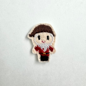 May include: Embroidered felt patch of a cartoon character wearing a brown hat, red shirt, and black trousers. The character has a white beard and a smiling face.