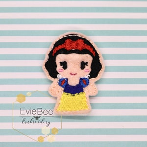 May include: A felt appliqué of Snow White, a Disney princess, with a red bow, yellow dress, blue bodice, and white collar. Her eyes are closed and she has a sweet smile.