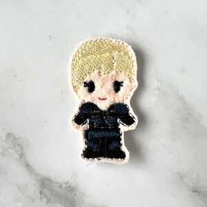 Op de afbeelding: Gestikt vilt patch van een cartoonfiguur met blond haar dat een zwarte jumpsuit draagt.