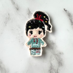 May include: Embroidered felt patch of a cartoon girl with black hair in a ponytail, wearing a blue and green outfit with a red belt. The girl has a pink face and big eyes.