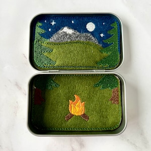 May include: A small metal tin with a felt scene of a mountain range under a starry night sky with a full moon. The tin opens to reveal a felt scene of a campfire in a forest.