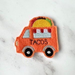 May include: Orange felt taco truck with a yellow taco on top. The truck has black wheels and the word "TACOS" embroidered on the side.